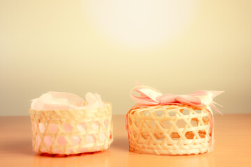 Fototapeta na wymiar A woven wooden basket paired with a pink bow-tied lid placed in the center of the warm, white background on a wooden floor