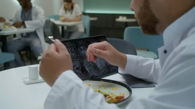 Professional doctor watches image of MRI or CT scan using digital tablet in clinic cafe. Medic eats his dinner, examines brain scanning results of patient. Medical staff have meal in hospital canteen.