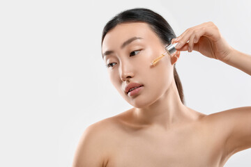 Beautiful young woman applying cosmetic serum onto her face on white background, space for text