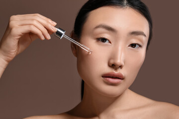 Fototapeta premium Beautiful young woman applying cosmetic serum onto her face on brown background, closeup