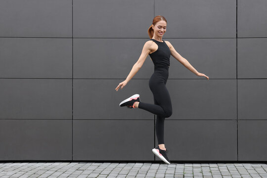 Beautiful woman in gym clothes jumping near dark grey wall on street