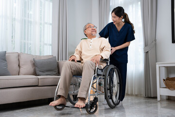 Fototapeta na wymiar A young Asian female nurse or physical therapist in a blue uniform, standing and talking happily with a senior Asian man sitting on a wheelchair while pushing him to relax in the living room at home.