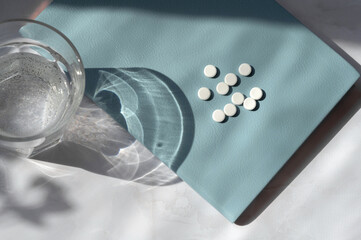 Minimal medicine concept, White pills on blue notebook, glass with water on white table background with sunlight shadows
