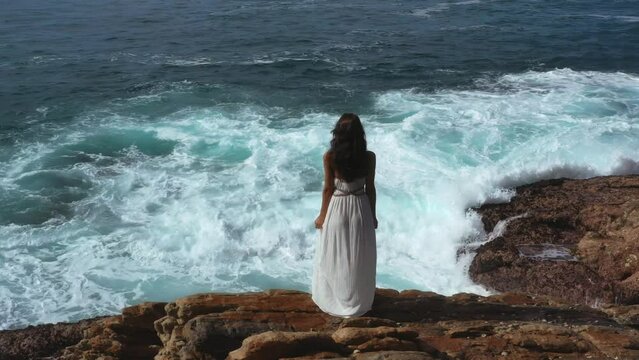 Aerial drone view of a beautiful bride in an amazing white dress standing on a cliff and looking at the sea. An epic scene with stormy waves and a gorgeous woman model. Professional 4k footage