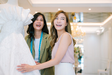 Fototapeta premium Asian bride is smiling while choosing wedding dress in modern wedding salon, Attractive designer girl using tape meter fitting on wedding dress at wedding studio