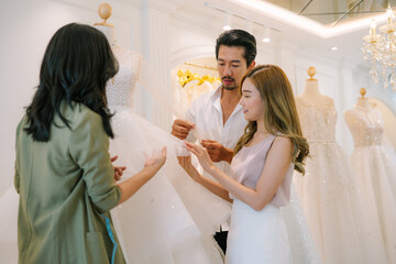 Naklejka premium Asian bride is smiling while choosing wedding dress in modern wedding salon, Attractive designer girl using tape meter fitting on wedding dress at wedding studio
