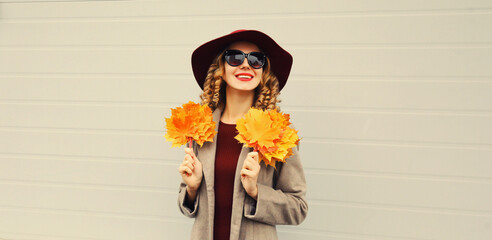 Autumn color style outfit, beautiful elegant smiling woman with yellow maple leaves wearing hat and coat jacket posing on gray background
