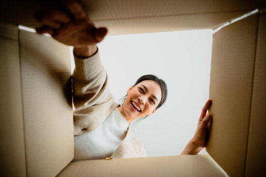 Surprised Young Asian Woman Unpacking. Opening Carton Box And Looking Inside. Packaging Box, Delivery Service. Human Emotions And Facial Expression