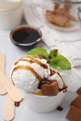 Scoops of tasty ice cream with caramel sauce, mint leaves and candies on white table, closeup