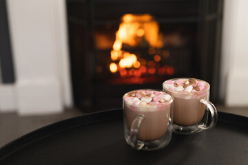 Close up of two cups of hot chocolate with marshmallows and open fire in fireplace, copy space