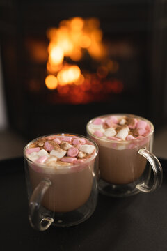 Close up of two cups of hot chocolate with marshmallows and open fire in fireplace, copy space