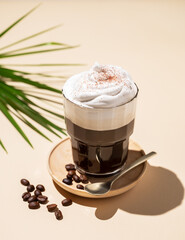 Iced coffee in a glass with foam on a yellow background with coffee beans, palm leaf and  shadows. Summer refreshment concept.