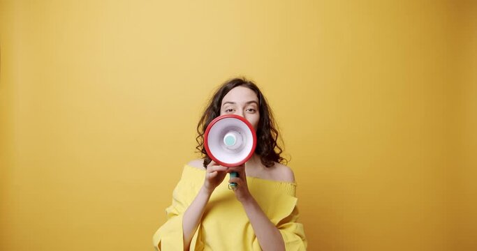 Happy brunette with megaphone making announcement. Static shot of cheerful young woman with dark hair gesticulating and smiling while using loudspeaker to make announcement against yellow background