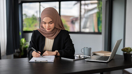 A focused Asian Muslim businesswoman is filling out forms or signing documents at her desk.
