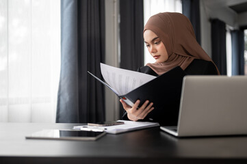 A professional Asian Muslim businesswoman focuses on examining business reports in a binder.