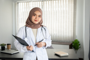 A professional Asian Muslim female doctor in a uniform and hijab stands in an office at a hospital.