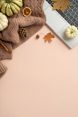 Autumn-infused workspace design. Vertical top view of laptop, cozy scarf, pattypans, acorn, cinnamon sticks, maple leaves, pine cone, dried orange slice on pastel beige backdrop. Space for text or ads