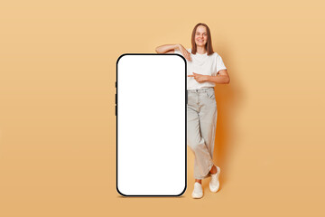 Smartphone display mockup. Internet shopping store. Cheerful happy brown haired adult woman indicating at big phone with white blank screen isolated over beige background