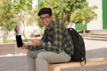 Young Indian middle class young man sitting at public place showing blank screen of smartphone