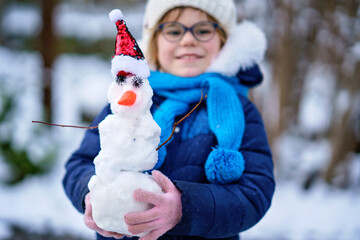 Cute little preschool girl with glasses making mini snowman. Adorable healthy happy child playing and having fun with snow, outdoors on cold day. Active leisure with children in winter