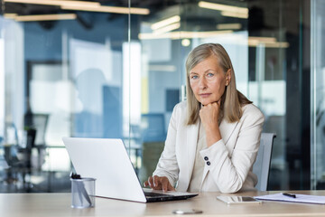Portrait of serious mature thinking businesswoman, gray haired female boss looking at camera, at workplace inside office, financier with laptop, confident accountant investor.