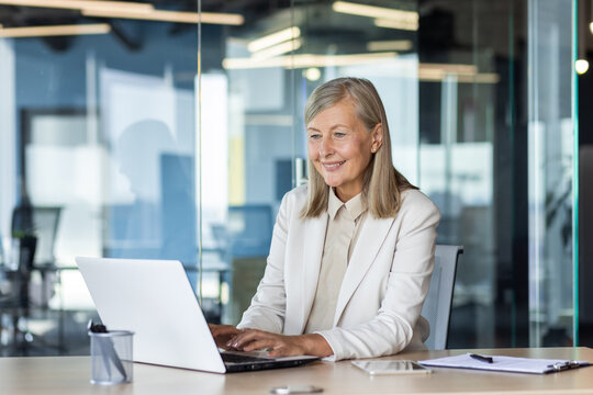 Senior mature gray-haired woman working at laptop inside office, boss businesswoman satisfied with achievement results typing on computer keyboard, boss in business suit.