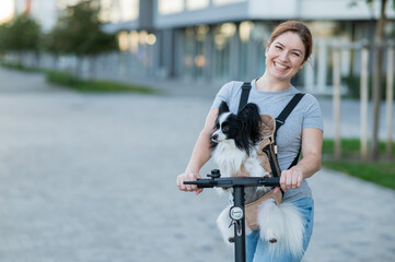 A woman rides an electric scooter with a dog in a backpack. Pappilion Spaniel Continental in a...