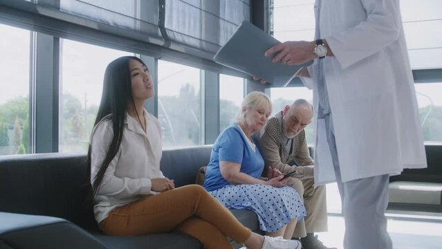Diverse people sit on couch in clinic lobby area, wait for doctors appointment. Doctor talks to Asian woman about medical examination, test results. Waiting room in modern medical center. Healthcare.