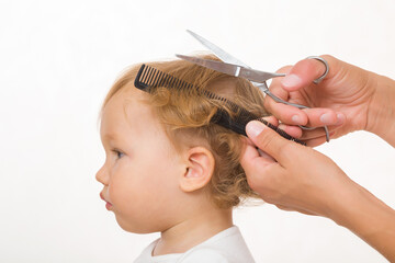 Young adult mother cutting baby boy blond wavy hair isolated on light gray background. Hands holding and using comb and scissors. Closeup. Side view.