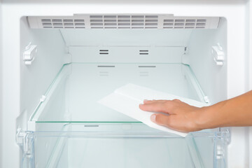 Young adult woman hand wiping glass shelf of refrigerator with dry white paper napkin. Closeup. Front view.