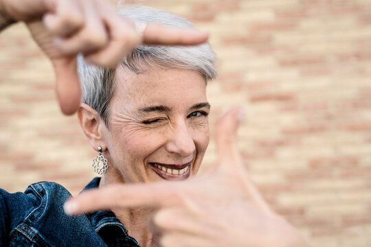 Smiling woman winking and showing finger frame sign in front of wall