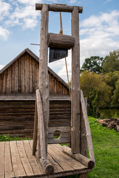 A guillotine on a wooden platform, behind it a river and a wooden barn.