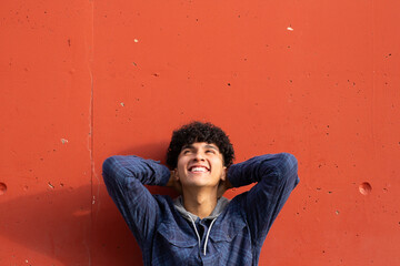 happy young guy standing by red wall with hands behind head and looking up