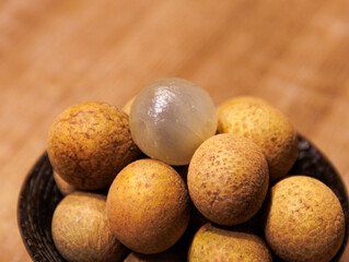 This is a close-up photo of some longan placed in a bowl. Most of the longan are brown and yellow, and the skin of the topmost longan has been peeled off.