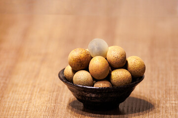 This is a close-up photo of some longan placed in a bowl. Most of the longan are brown and yellow, and the skin of the topmost longan has been peeled off.