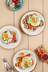 Aesthetic composition of Viennese waffles, adorned with toppings of egg and salmon, egg and bacon, and egg with meat. Served on a beige wooden background with utensils and fresh ingredients