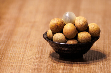 This is a close-up photo of some longan placed in a bowl. Most of the longan are brown and yellow,...