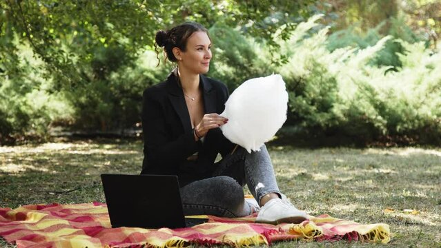 A business woman in a suit spends time outdoors in a city park on the grass, and eats cotton candy. Positive, carefree young woman rejoices after receiving a message on her laptop. Conclusion of a dea