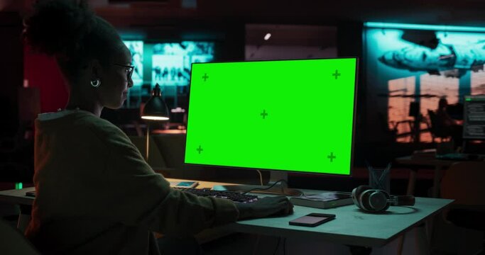 Portrait of Creative Black Woman Sitting at Her Desk Using Desktop Computer with Mock-up Green Screen. Female Specialist Working at Night in Game Development Startup, Finishing Details on a Project