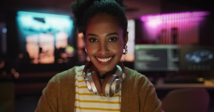 Close Up Portrait of a Young Black Woman with Headphones, Looking at Camera, and Smiling in a Creative Office. Beautiful Diverse Female Working for her Dream Agency, Feeling Empowered