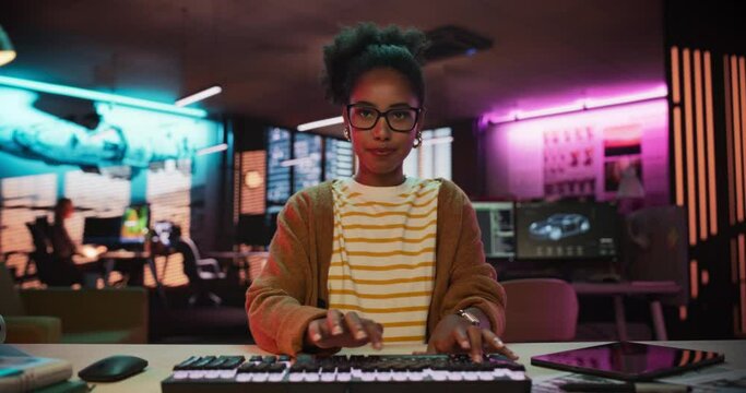 Portrait of a Black Female Developer Writing Code in a Creative Office with Neon Lights. Young Woman Focused on Typing on Keyboard and Working on Desktop Computer. Point of View of Screen Monitor