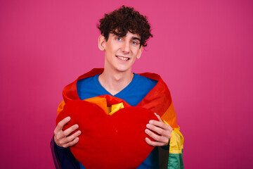 Young attractive curly man posing on a pink background with a plush heart and a gay flag.