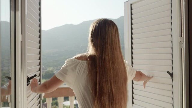 Young brunette, dressed elegantly, opens her window blinds, ventures to the terrace, and looks at the mountain sunrise or sunset a serene stock footage capturing vacation bliss.