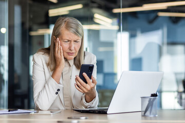 Mature adult business woman inside office at workplace received online notification message with bad news on phone, female boss with gray hair using phone frustrated and displeased reading online.