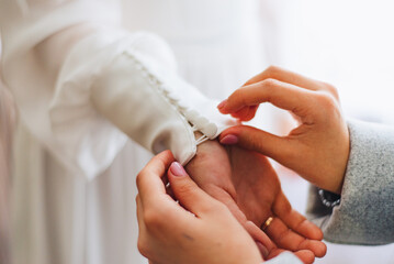 The bridesmaid fastens the buttons on the sleeve of the bride's dress. The bride straightens her...