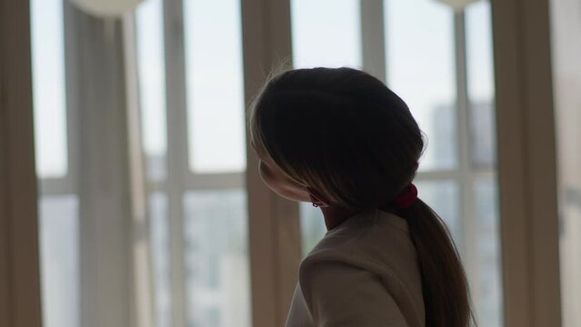 Side view tracking shot of athletic little child girl doing stretching exercises for shoulders and neck before fitness training at dark bedroom by window. Sporty female kid warming up muscles indoors.