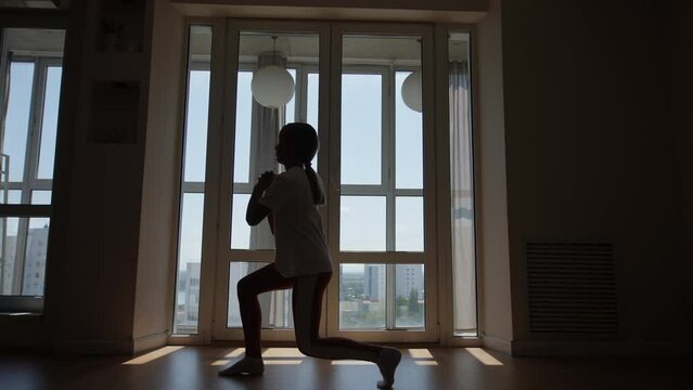 Wide side view shot of athletic little child girl having practice sport during home workout, doing squats training, maintain healthy lifestyle in dark room by window. Concept of exercises for children