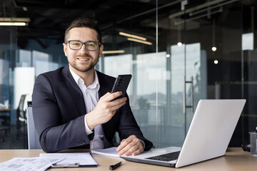 Portrait of a young successful businessman working in the office on a laptop and using a mobile phone. He looks at the camera with a smile