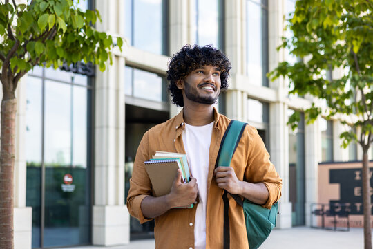 Portrait Of Young Indian Male Student, Applicant Standing Outside Campus, Holding Books And Backpack, Preparing For Exam. Smiling And Confidently Looking To The Side