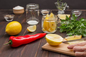 Hands chop a lemon on cutting board. Lemon in jar. Chili pepper, whole lemon and salt.